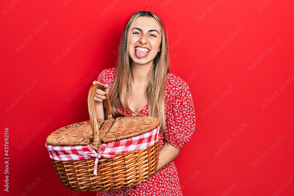 Sticker Beautiful hispanic woman holding picnic wicker basket sticking tongue out happy with funny expression.