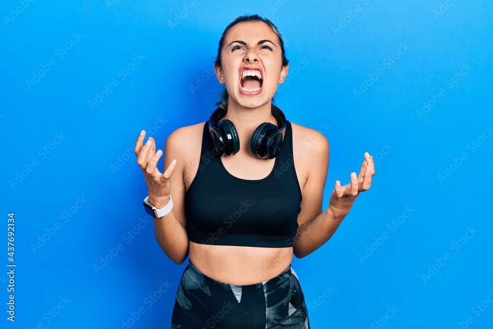 Sticker Beautiful hispanic woman wearing gym clothes and using headphones crazy and mad shouting and yelling with aggressive expression and arms raised. frustration concept.