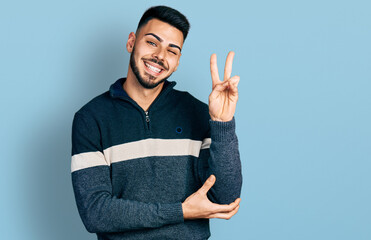 Young hispanic man with beard wearing casual winter sweater smiling with happy face winking at the camera doing victory sign. number two.
