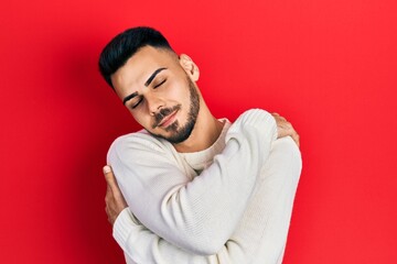 Young hispanic man with beard wearing casual winter sweater hugging oneself happy and positive, smiling confident. self love and self care