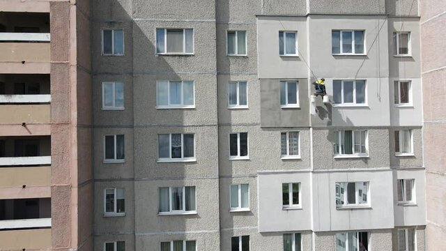 An industrial climber suspended on ropes and paints the wall of a building with a roller. A male worker performs work on the insulation of the facade of a multi-storey building. View from the drone.