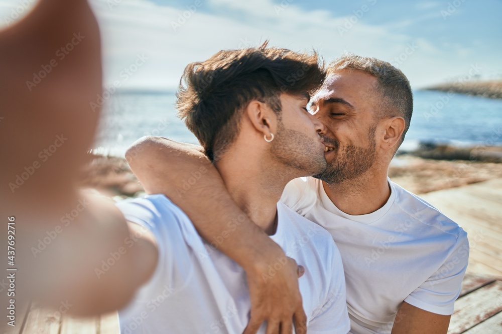 Poster young gay couple kissing making selfie by the camera at the beach.