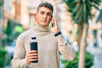 Young hispanic man with serious expression talking on the smartphone and holding bottle of water at the city.