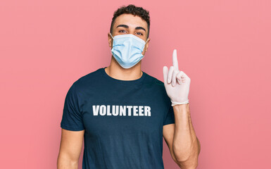 Hispanic young man wearing medical mask and volunteer t shirt pointing finger up with successful idea. exited and happy. number one.