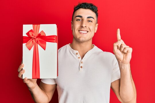 Hispanic young man holding gift smiling with an idea or question pointing finger with happy face, number one