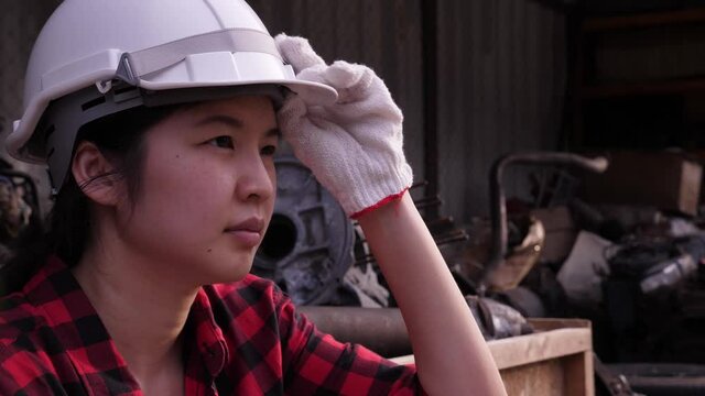 Side view portrait of depressed Asian woman wearing lumberjack shirt, sitting, looking away, being tired and abstracted, taking off safety hard hat. Female worker or engineer concept.