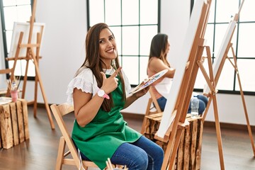 Fototapeta premium Young hispanic artist women painting on canvas at art studio approving doing positive gesture with hand, thumbs up smiling and happy for success. winner gesture.