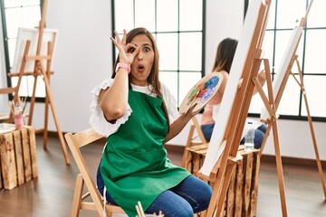 Young hispanic artist women painting on canvas at art studio doing ok gesture shocked with surprised face, eye looking through fingers. unbelieving expression.