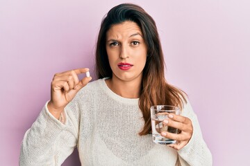 Beautiful hispanic woman holding pill and glass of water skeptic and nervous, frowning upset...