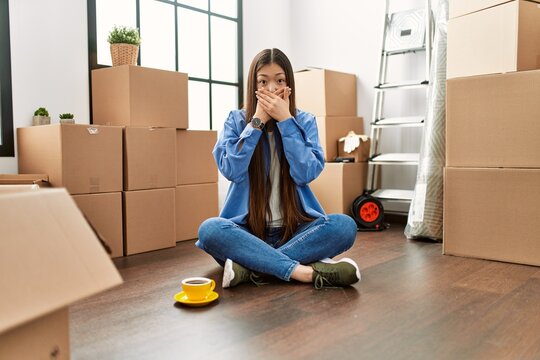 Young chinese girl sitting on the floor at new home shocked covering mouth with hands for mistake. secret concept.