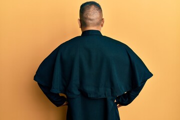 Obraz premium Young latin priest man standing over yellow background standing backwards looking away with arms on body