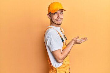 Hispanic young man wearing handyman uniform pointing aside with hands open palms showing copy space, presenting advertisement smiling excited happy