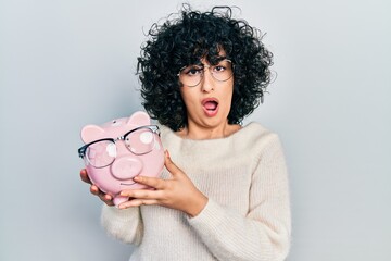 Young middle east woman holding piggy bank with glasses in shock face, looking skeptical and sarcastic, surprised with open mouth