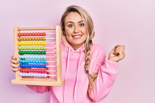 Beautiful young blonde woman holding traditional abacus screaming proud, celebrating victory and success very excited with raised arm