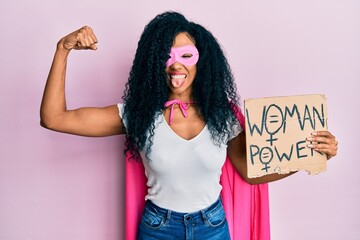 Middle age african american woman wearing super hero costume holding woman power banner sticking tongue out happy with funny expression.