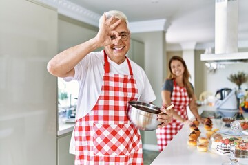 Middle age hispanic couple wearing apron cooking homemade pastry smiling happy doing ok sign with hand on eye looking through fingers