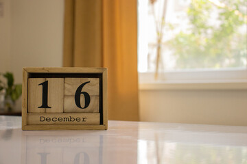 Wooden blocks of the calendar represents the date 16 and the month of December on the background of a window, curtain and a plant