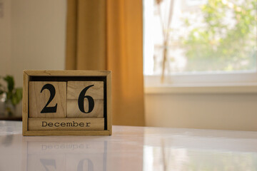 Wooden blocks of the calendar represents the date 26 and the month of December on the background of a window, curtain and a plant