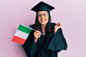 Young hispanic woman wearing graduation uniform holding italy flag screaming proud, celebrating victory and success very excited with raised arm