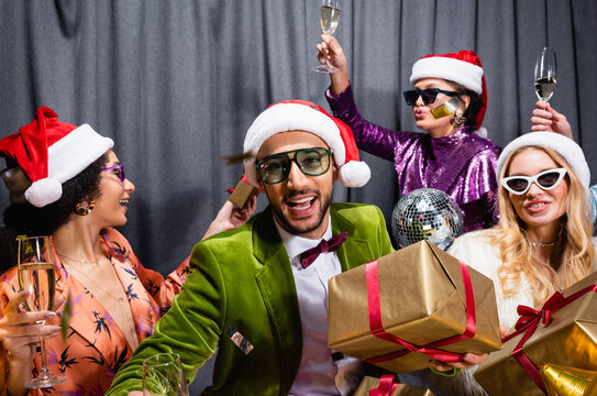 smiling interracial friends in santa hats drinking champagne and celebrating new year on grey background.
