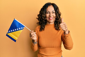 Middle age hispanic woman holding bosnia herzegovina flag smiling happy and positive, thumb up doing excellent and approval sign