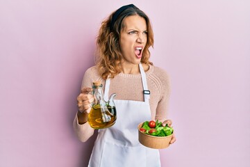 Young caucasian woman wearing apron holding olive oil can and salad angry and mad screaming frustrated and furious, shouting with anger. rage and aggressive concept.