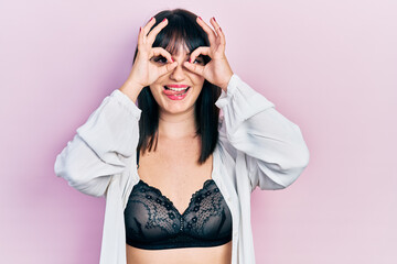 Young hispanic woman wearing lingerie doing ok gesture like binoculars sticking tongue out, eyes looking through fingers. crazy expression.