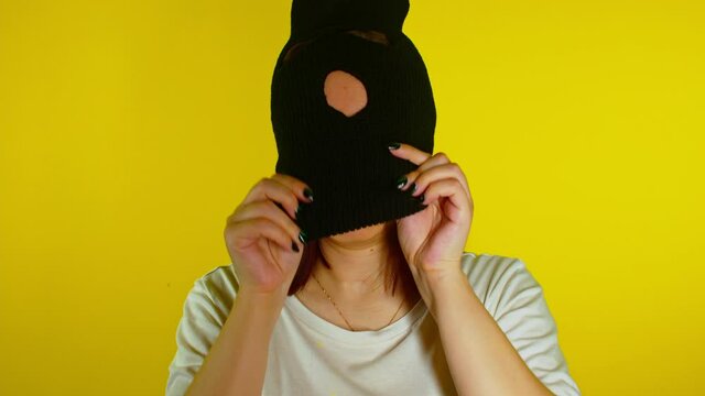 Close up young woman puts on black balaclava on yellow background. Secretive female puts on mask, looking at camera.