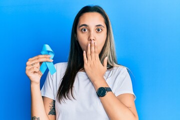 Young hispanic woman holding blue ribbon covering mouth with hand, shocked and afraid for mistake. surprised expression