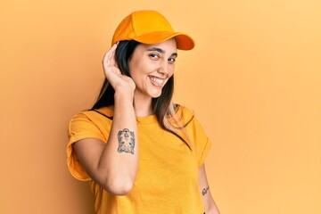 Young hispanic woman wearing delivery uniform and cap smiling with hand over ear listening an hearing to rumor or gossip. deafness concept.