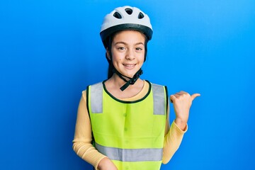 Beautiful brunette little girl wearing bike helmet and reflective vest smiling with happy face looking and pointing to the side with thumb up.