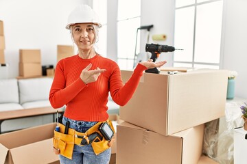 Middle age grey-haired woman wearing hardhat standing at new home amazed and smiling to the camera while presenting with hand and pointing with finger.