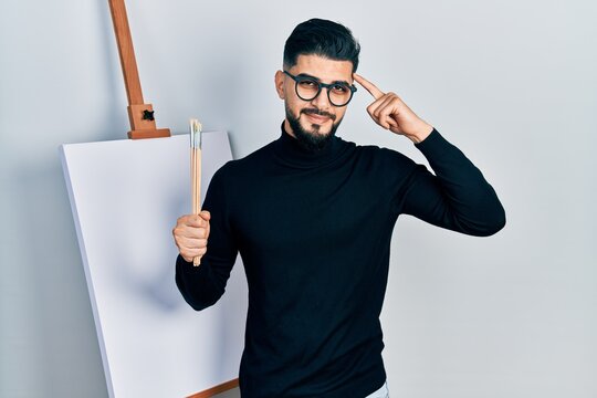 Handsome man with beard holding brushes close to easel stand smiling pointing to head with one finger, great idea or thought, good memory