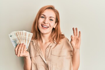 Young caucasian woman holding 100 danish krone banknotes doing ok sign with fingers, smiling friendly gesturing excellent symbol
