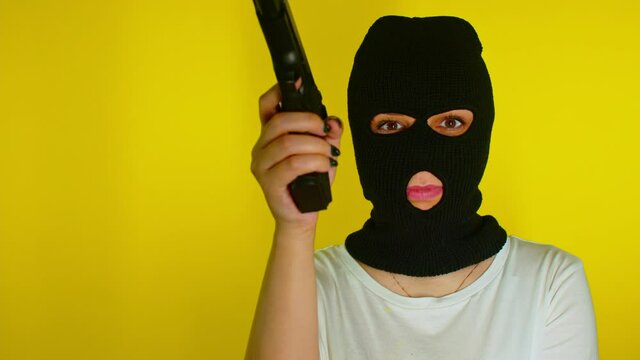 Unrecognizable woman in black balaclava with gun on yellow background. Dangerous female in mask takes aim at camera, shoots and laughs. Slow motion