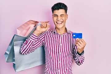Young hispanic man holding shopping bags and credit card winking looking at the camera with sexy...