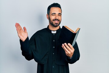Handsome hispanic priest man with beard holding bible and christian cross winking looking at the...