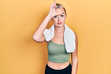 Young blonde girl wearing sportswear and towel making fun of people with fingers on forehead doing loser gesture mocking and insulting.