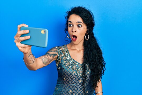 Young woman wearing bindi and traditional kurta dress taking a selfie photo with smartphone scared and amazed with open mouth for surprise, disbelief face