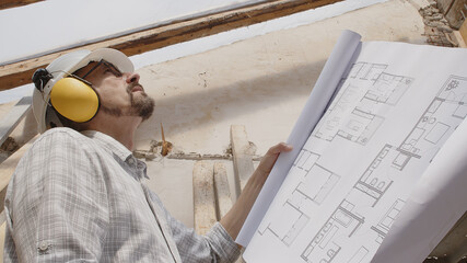 architect man at work, wearing helmet and headphones looking at blueprint , check the construction...