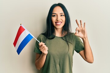 Young hispanic girl holding holland flag doing ok sign with fingers, smiling friendly gesturing excellent symbol - obrazy, fototapety, plakaty