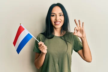 Fotobehang Young hispanic girl holding holland flag doing ok sign with fingers, smiling friendly gesturing excellent symbol © Krakenimages.com