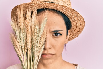 Young hispanic girl wearing summer hat holding spike wheat over eye skeptic and nervous, frowning...