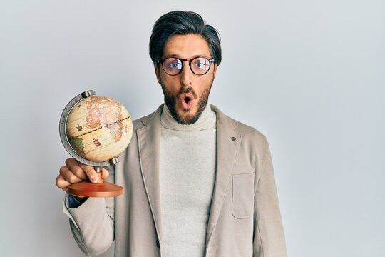 Young hispanic man holding vintage world ball scared and amazed with open mouth for surprise, disbelief face