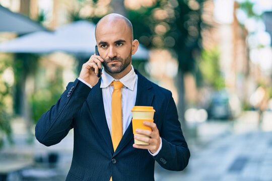 Young hispanic bald businessman with serious expression talking on the smartphone drinking coffee at the city