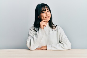 Young brunette woman with bangs wearing casual clothes sitting on the table smiling looking...