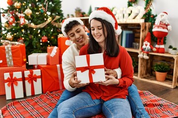 Obraz na płótnie Canvas Young hispanic couple smiling happy and hugging. Sitting on the floor wearing christmas hat holding gift at home.