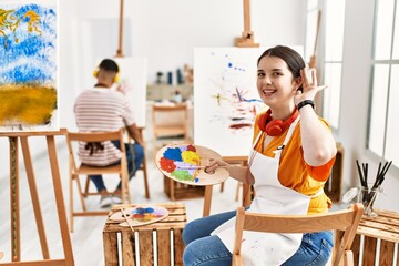 Young artist woman painting on canvas at art studio smiling with hand over ear listening an hearing...