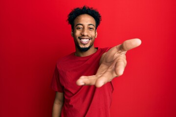 Young african american man with beard wearing casual red t shirt smiling friendly offering handshake as greeting and welcoming. successful business.