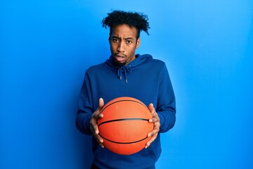 Young african american man with beard holding basketball ball in shock face, looking skeptical and sarcastic, surprised with open mouth